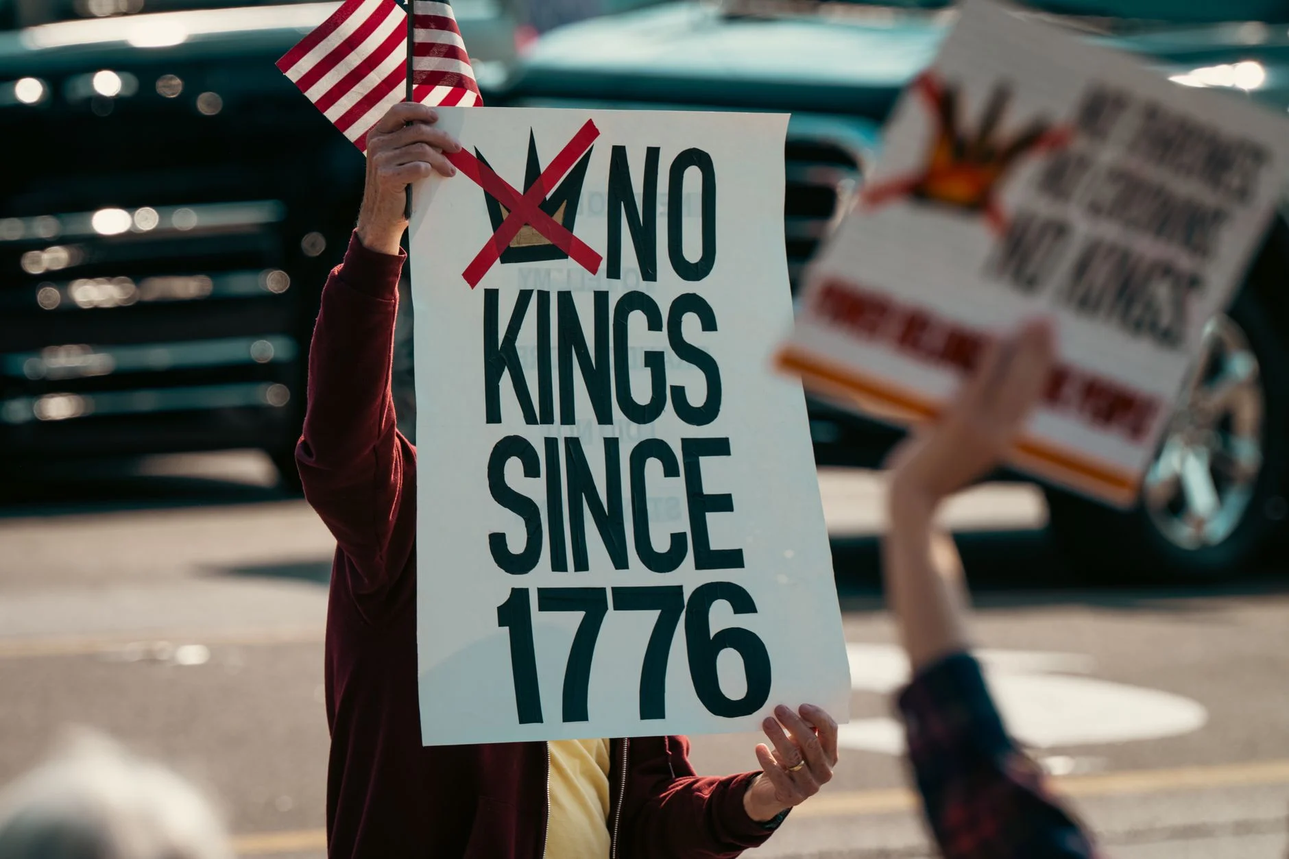 Protest sign displaying 'No Kings Since 1776' held alongside a small US flag, emphasizing democratic sentiments.