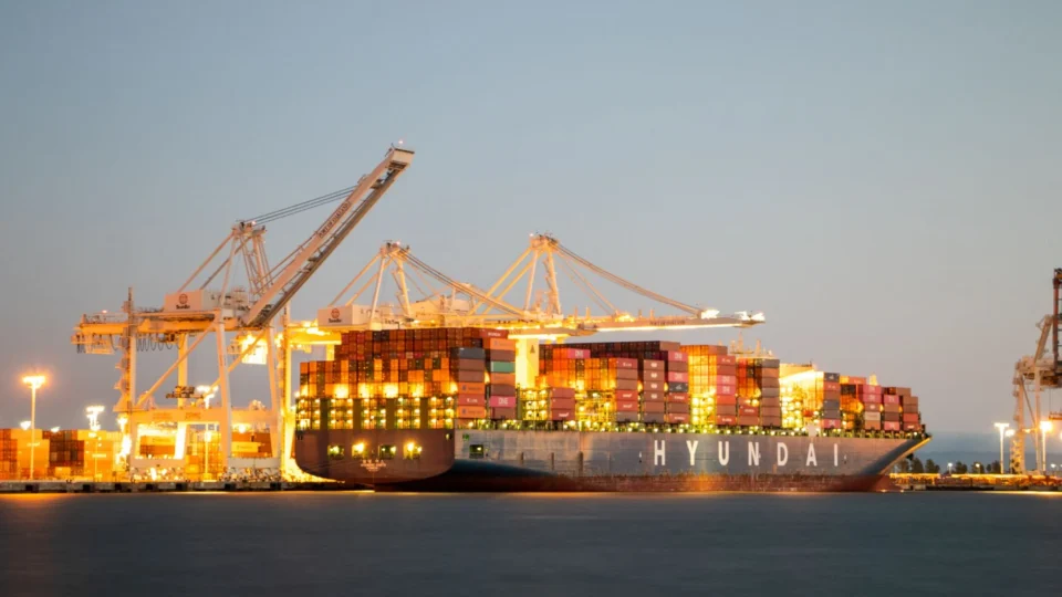 Container ship docked at port under cranes during twilight, illuminated against the evening sky.