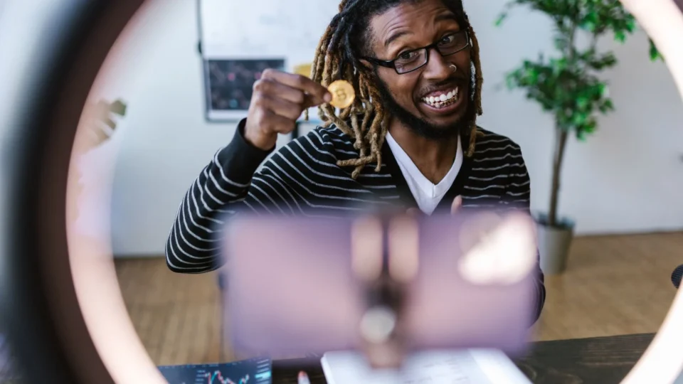 African American man smiling while holding a Bitcoin in front of a smartphone for a video recording.