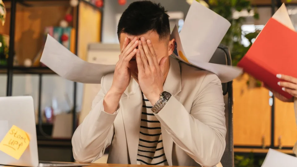 Stressed businessman overwhelmed by paperwork in office environment, demonstrating burnout.