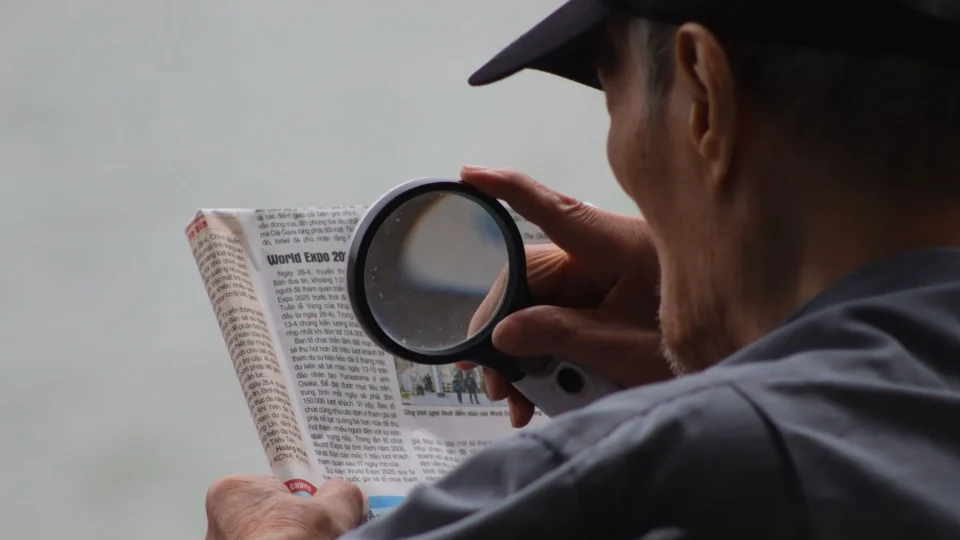 Elderly man using a magnifying glass to enhance text in a newspaper.