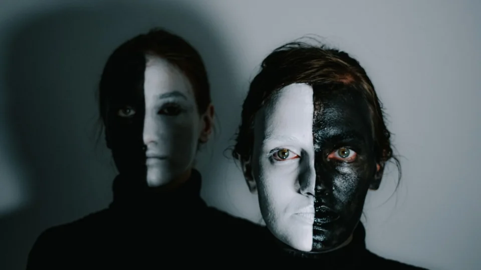 Unemotional models with face painted in white and black paints standing in room with light wall and looking at camera