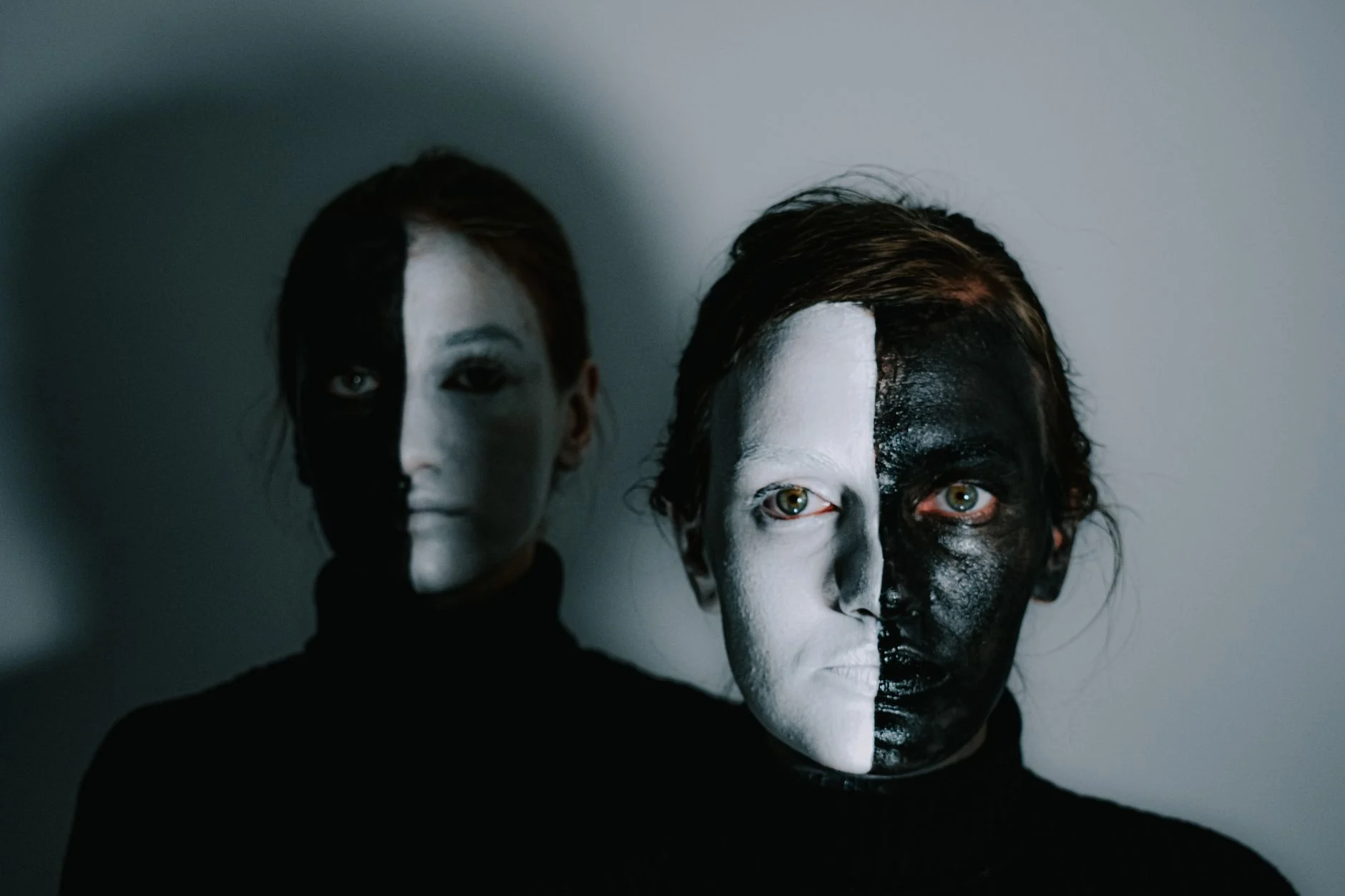 Unemotional models with face painted in white and black paints standing in room with light wall and looking at camera