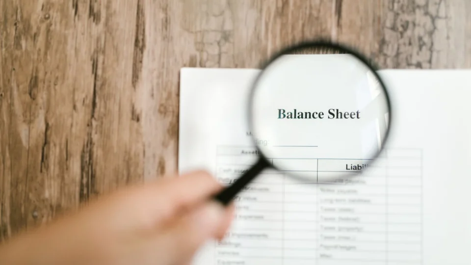 Close-up of a balance sheet document on wooden surface with a magnifying glass held by a hand.