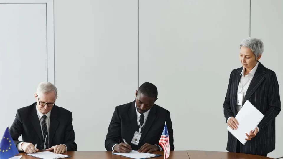 Executives signing international agreement with EU and US flags displayed on a wooden table.