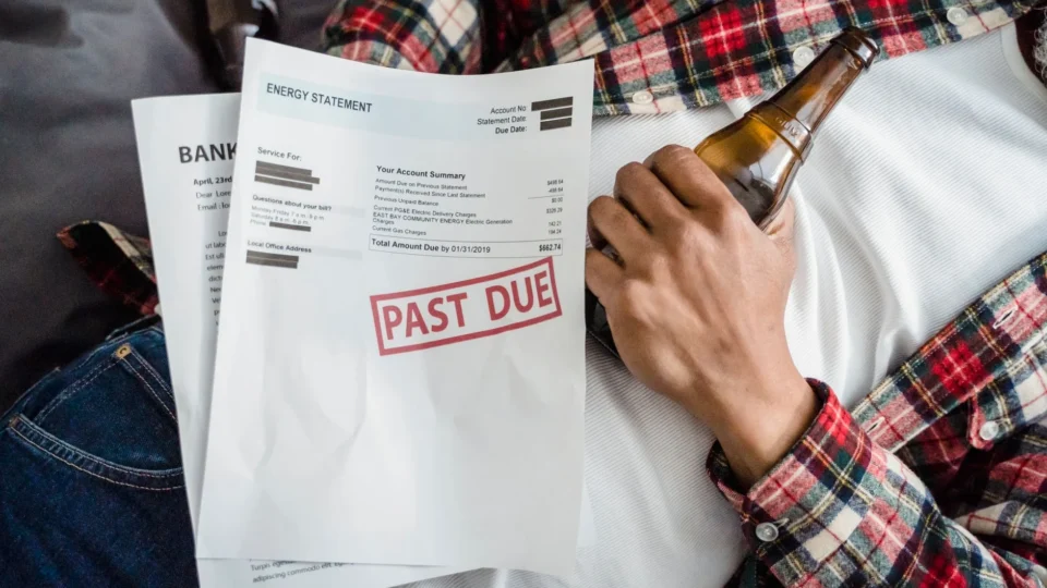 A man relaxes with a beer bottle while holding past due bills, highlighting financial stress.