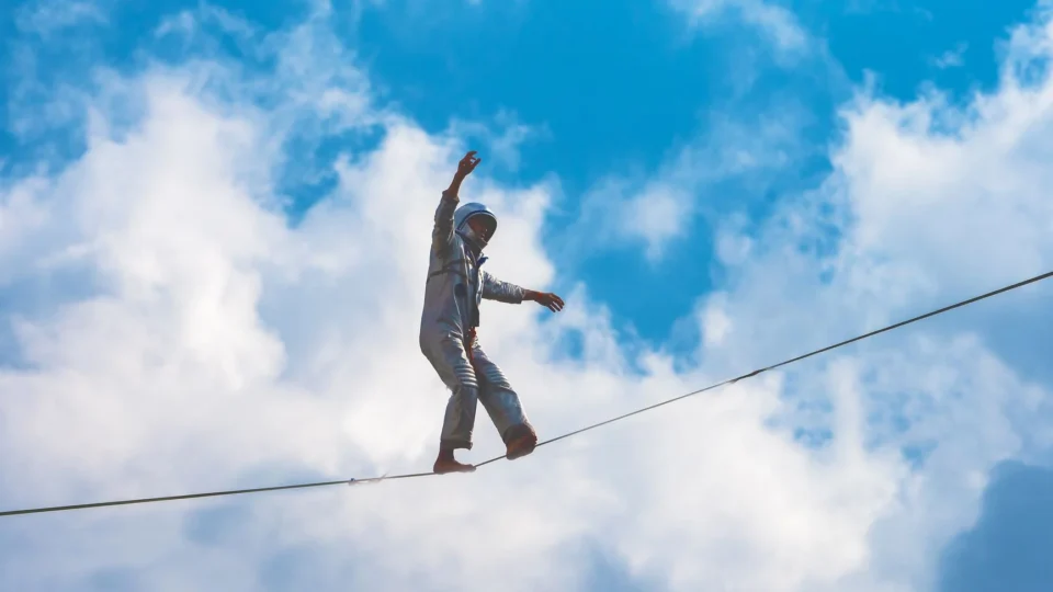 A performer balances on a high wire against a bright blue sky and clouds.