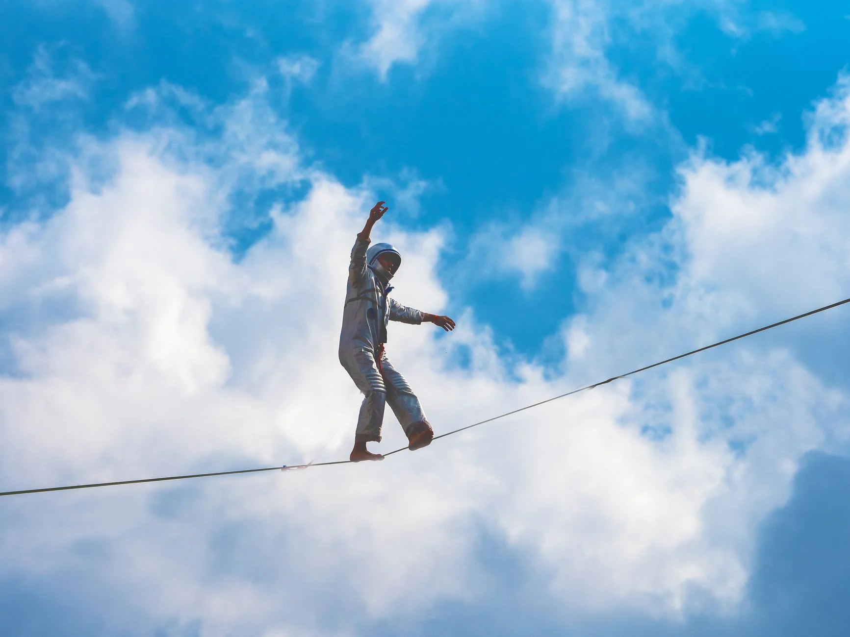 A performer balances on a high wire against a bright blue sky and clouds.