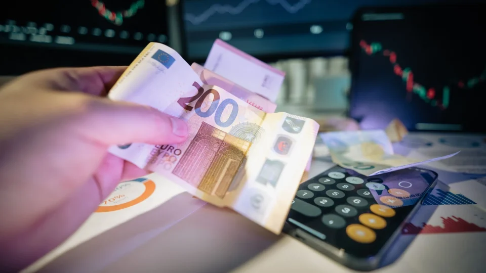 A hand holds euro notes with financial charts and a calculator, symbolizing investment and trading.
