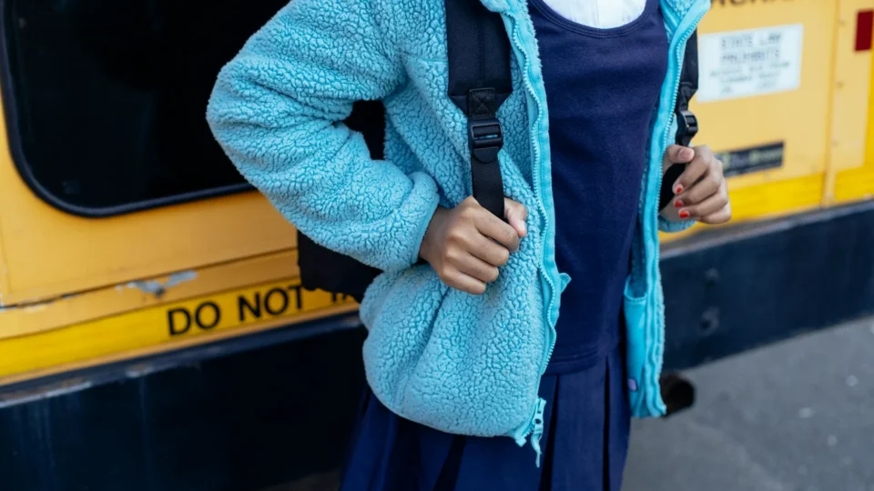 Crop anonymous ethnic schoolgirl in uniform with rucksack standing on roadway near bus in town