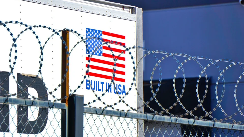 Industrial scene with barbed wire fence and American flag logo on building.