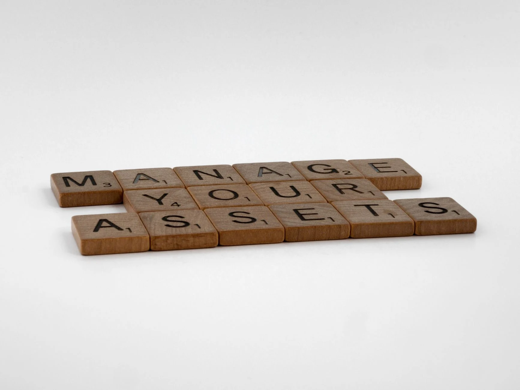 Wooden Scrabble tiles spelling 'Manage Your Assets' on a white background.