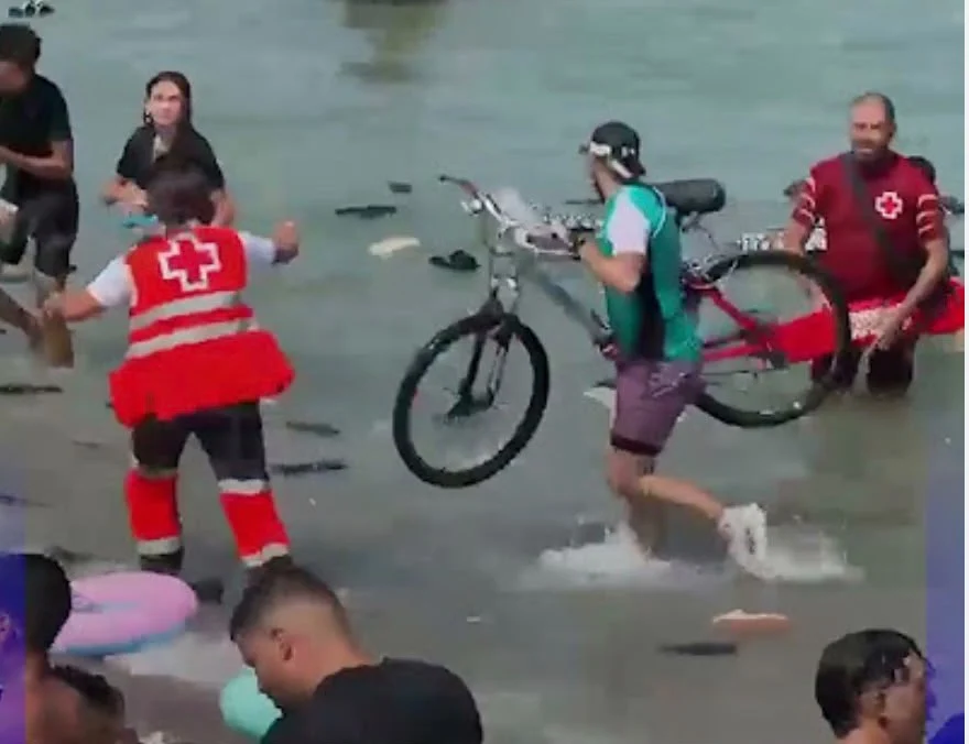 A migrant carrying a bicycle through shallow water as Red Cross workers assist nearby