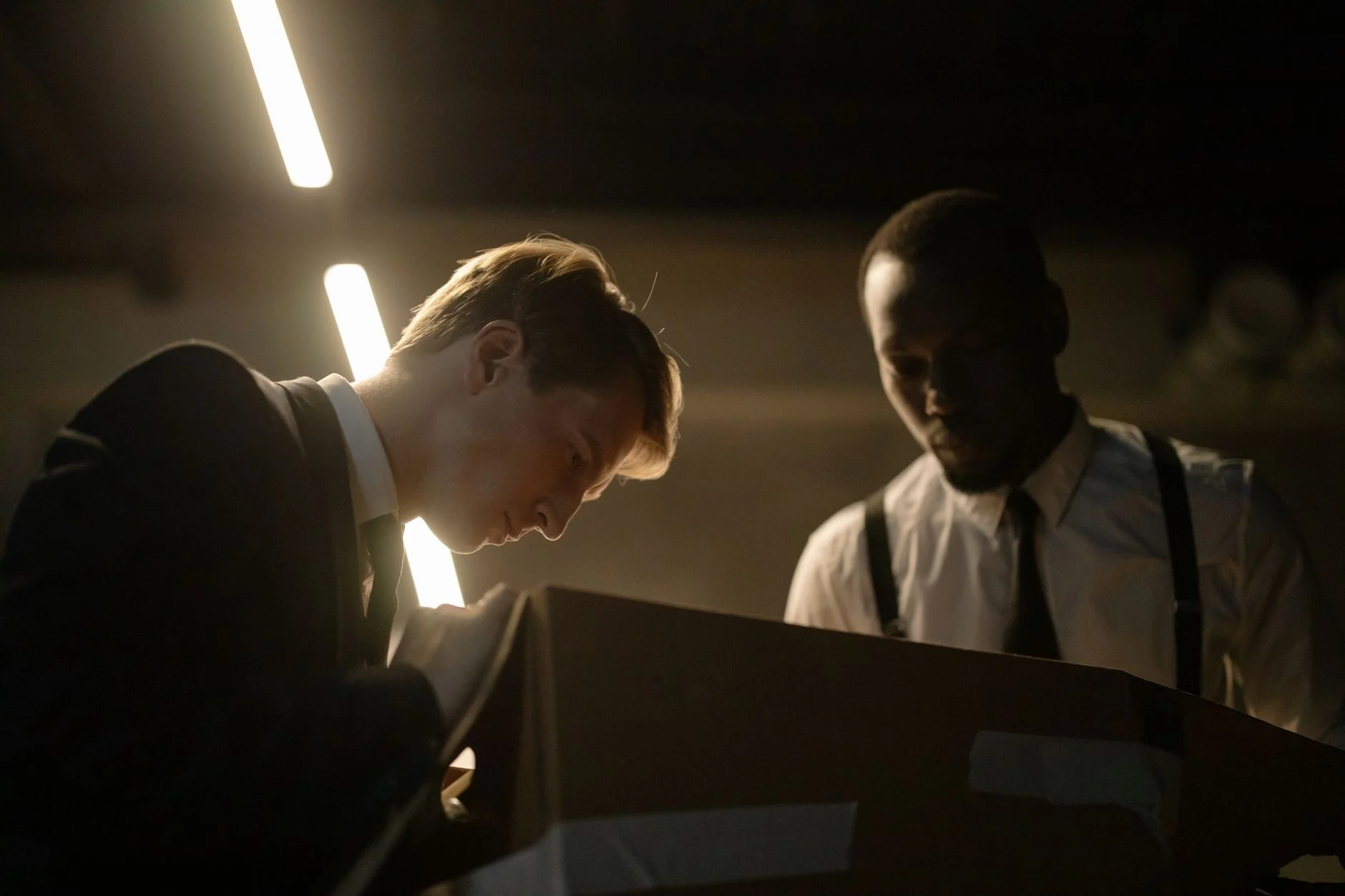 Two detectives examine a box in dimly lit room, focused on investigation.