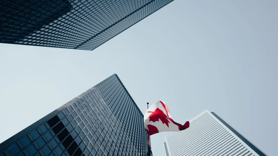 Dramatic low-angle view of skyscrapers and a Canadian flag in downtown Toronto, under a clear blue sky.