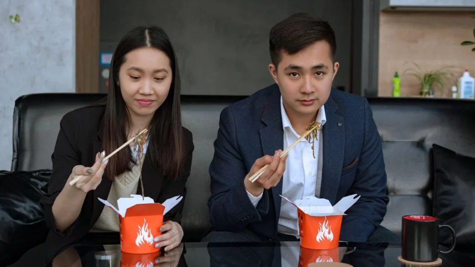 A young man and woman in business attire enjoying a casual lunch with chopsticks.