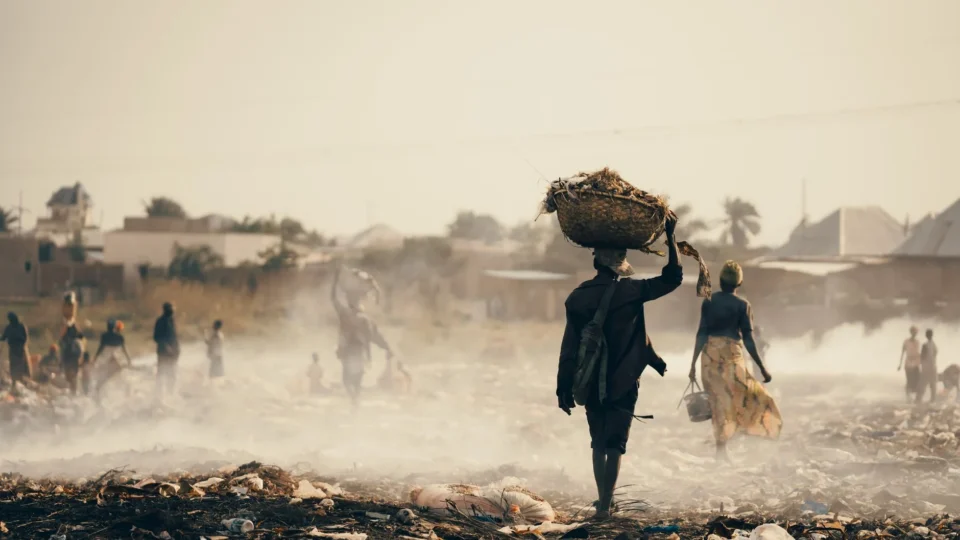 People walk through waste in a smoke-filled landscape in Burundi, highlighting pollution.