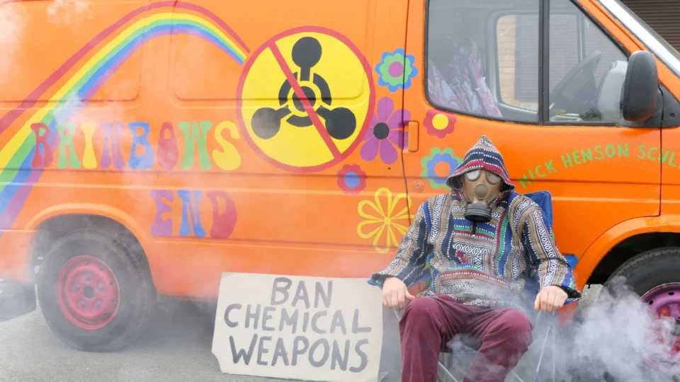 A protester in a gas mask sits by a colorfully painted van with an anti-chemical weapons sign.