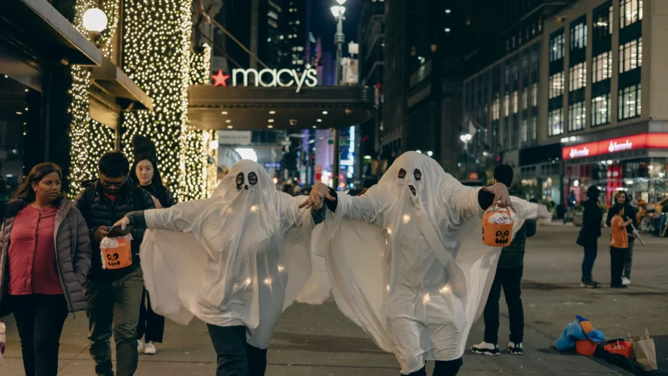 Adults in ghost costumes walk in New York City at night, celebrating Halloween.