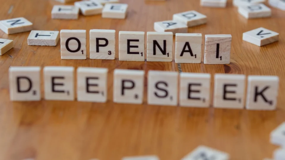 Close-up of wooden Scrabble tiles spelling OpenAI and DeepSeek on wooden table.