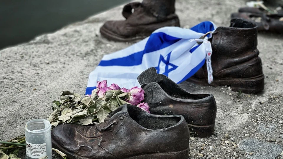 Sculpture of shoes with Israeli flag and flowers on riverbank, symbolizing remembrance.