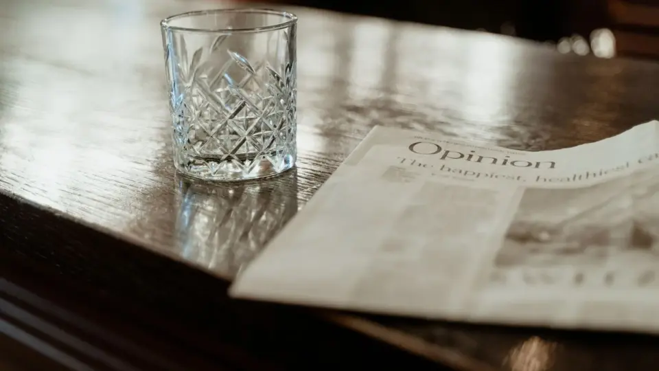 A close-up of a crystal drinking glass and newspaper on a polished wooden bar counter.