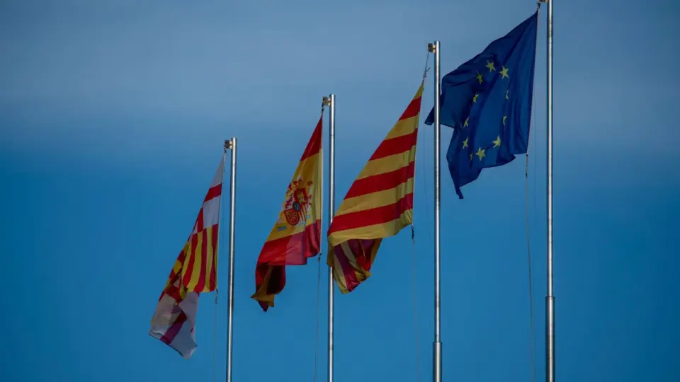 Flags of Spain, Catalonia, and Europe on flagpoles against a blue sky in Barcelona.